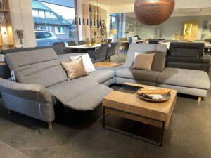 Zwei moderne graue Sofas mit unterschiedlichen Designs stehen in einer stilvollen Möbelausstellung einander gegenüber. Beide Sofas sind mit dekorativen Kissen ausgestattet. Dazwischen steht ein quadratischer Couchtisch mit Holzplatte und schwarzem Metallrahmen, darauf liegt eine Schale mit Holzdekoration. Im Hintergrund sind weitere Möbelstücke wie Tische, Stühle und Regale ausgestellt.