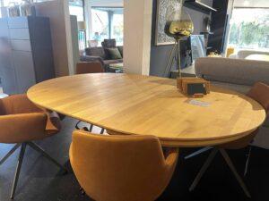 Runder Esstisch aus Holz mit vier gepolsterten Stühlen in warmem Cognac-Ton. Die Stühle haben Armlehnen und ein modernes Kreuzfuß-Gestell aus Metall. Der Tisch steht in einem Ausstellungsraum mit verschiedenen Wohnmöbeln, im Hintergrund sind ein graues Sofa, ein dunkelblauer Wandschrank und eine große Stehlampe mit Glas-Schirm zu sehen.