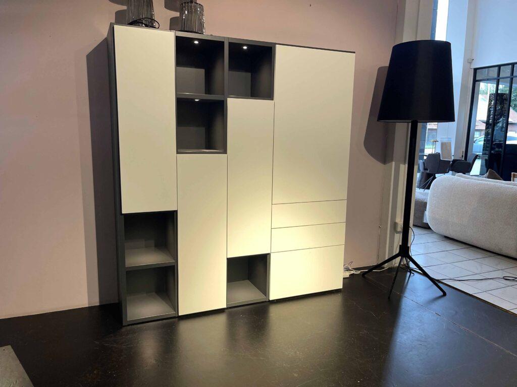 Modernes Sideboard in asymmetrischem Design mit geschlossenen, weißen Fronten und offenen, anthrazitfarbenen Fächern. Einige der offenen Fächer sind beleuchtet. Das Möbelstück steht vor einer hellrosafarbenen Wand auf dunklem Boden. Rechts daneben befindet sich eine große, schwarze Stehlampe mit klassischem Lampenschirm. Im Hintergrund ist ein heller Wohnbereich mit großen Fenstern zu sehen.